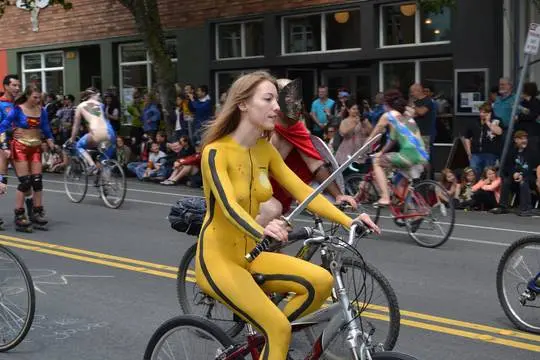 Slim brunette woman with yellow body paint riding a bicycle at a street fair, topless and nude