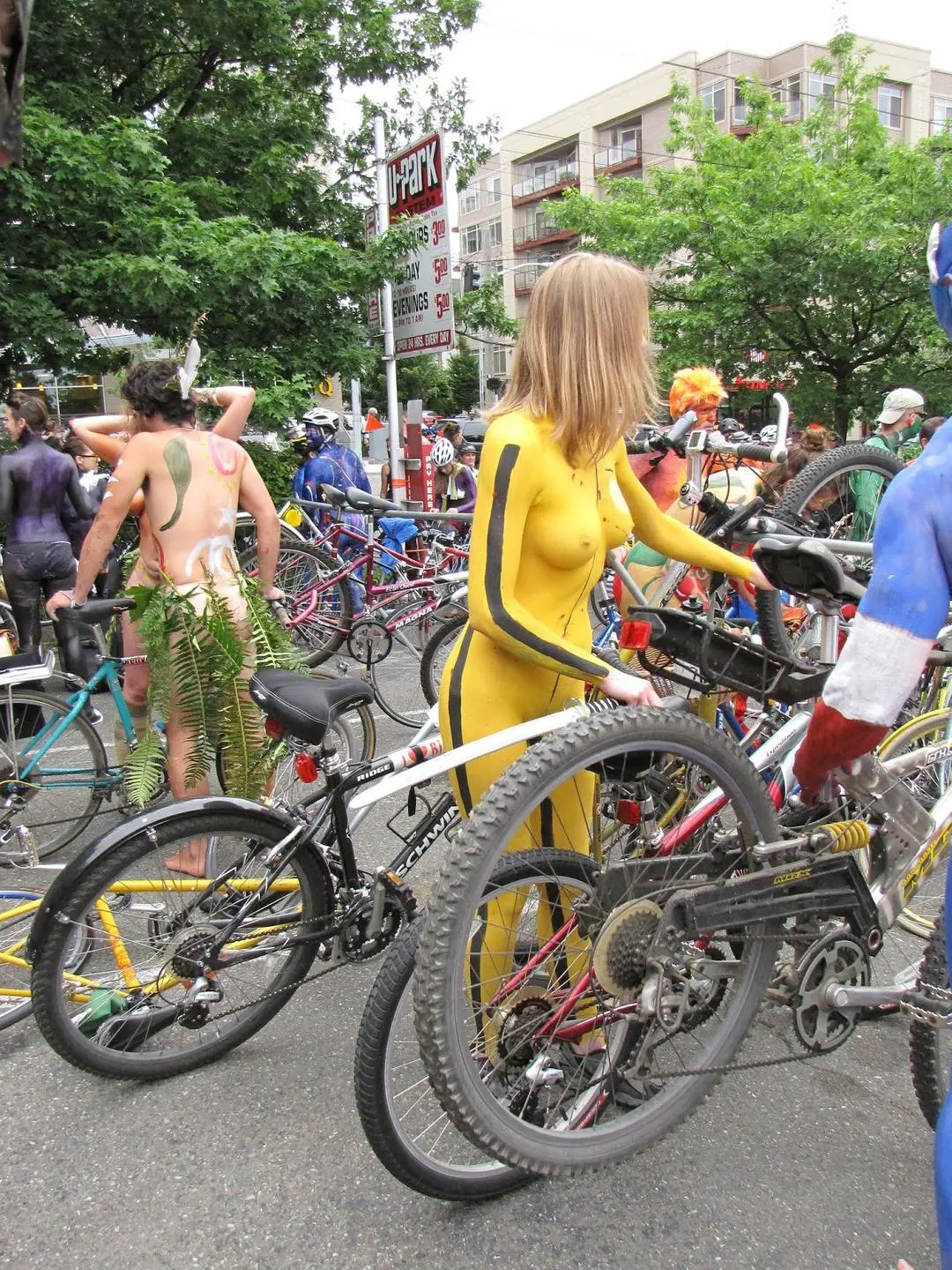 Slim brunette woman with yellow body paint riding a bicycle at a street fair, topless and nude