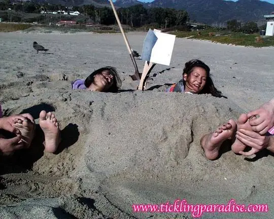 Asian women tickling feet and toes with a pink feather while buried in the sand at the beach