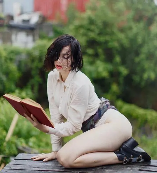 Bottomless librarian in a white shirt and plaid skirt reading a book while kneeling on a wooden garden table