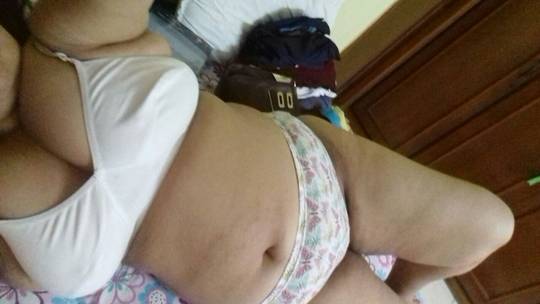 Curvy Indian bhabhi in white bra and patterned panties posing for a selfie in a bedroom.