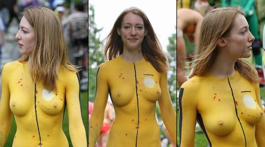 Slim brunette woman with yellow body paint riding a bicycle at a street fair, topless and nude