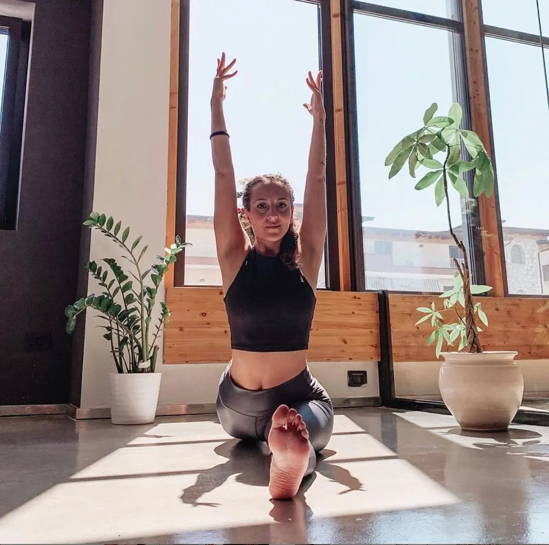 Athletic woman with brown hair in a black crop top and grey leggings doing yoga between potted plants