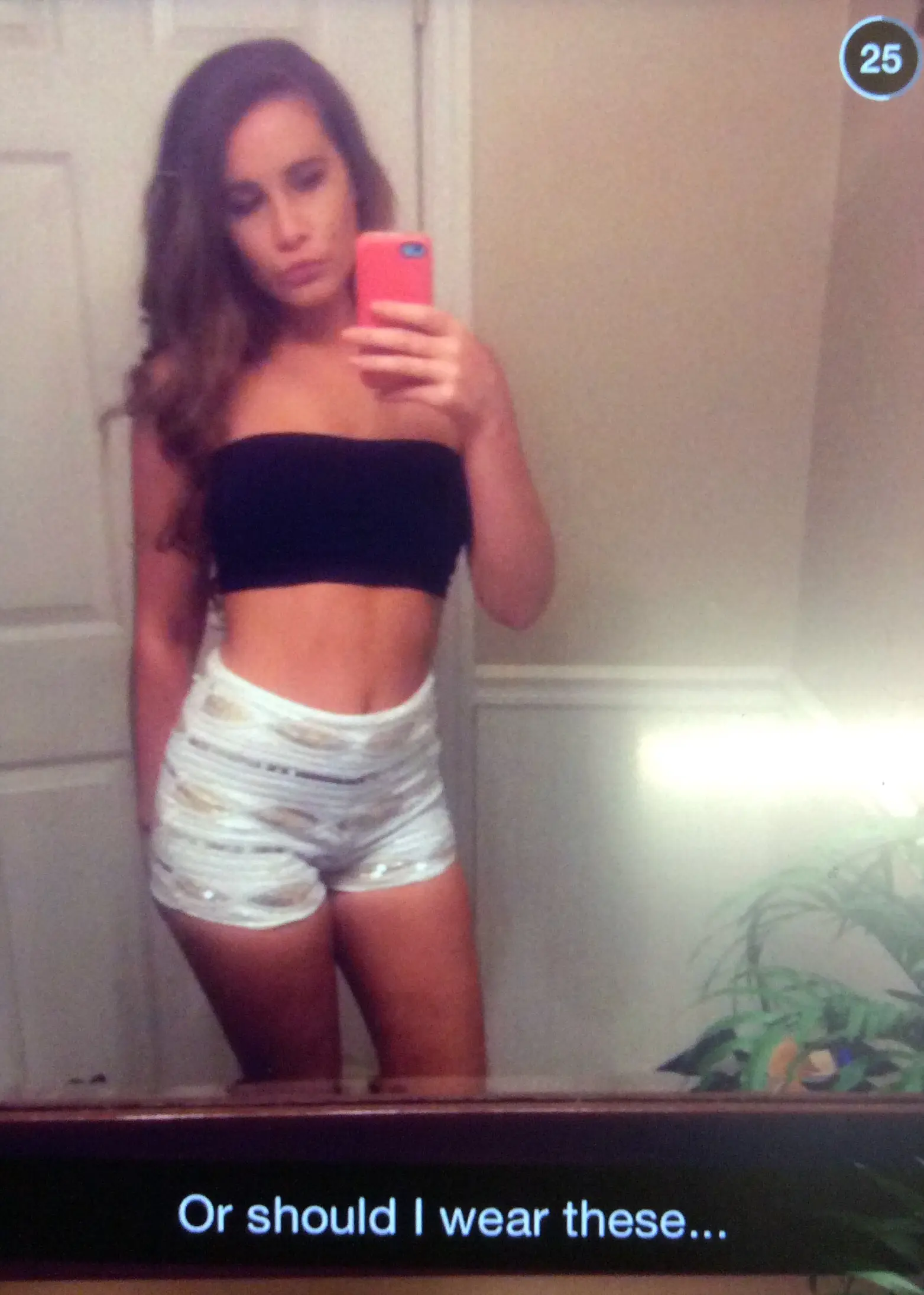 Slim brunette woman in a black tube top and white shorts posing for mirror selfies