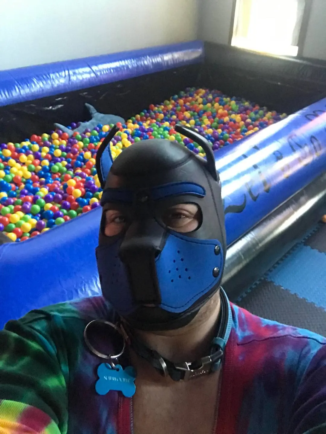 White man in a blue and black puppy play mask, collar, and tie-dye shirt taking a selfie by a ball pit