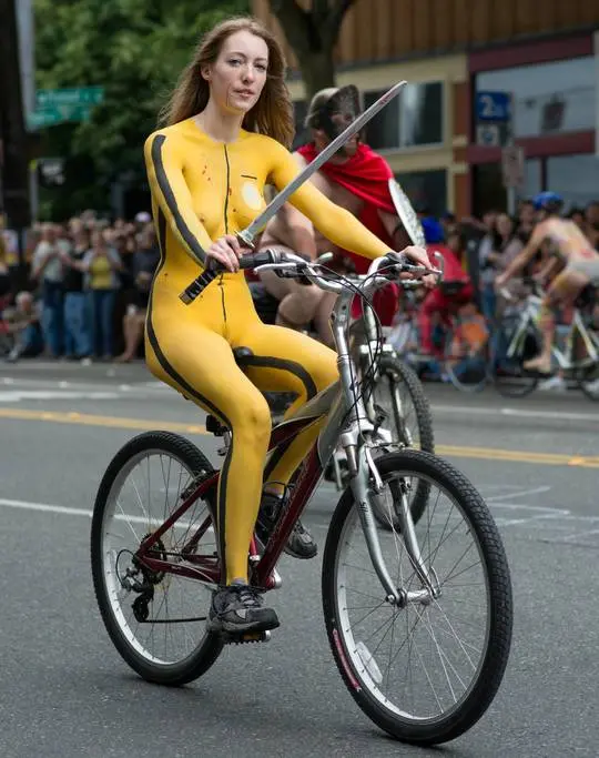 Slim brunette woman with yellow body paint riding a bicycle at a street fair, topless and nude