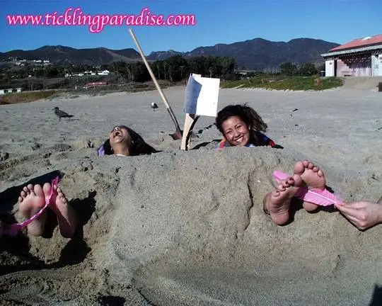 Asian women tickling feet and toes with a pink feather while buried in the sand at the beach