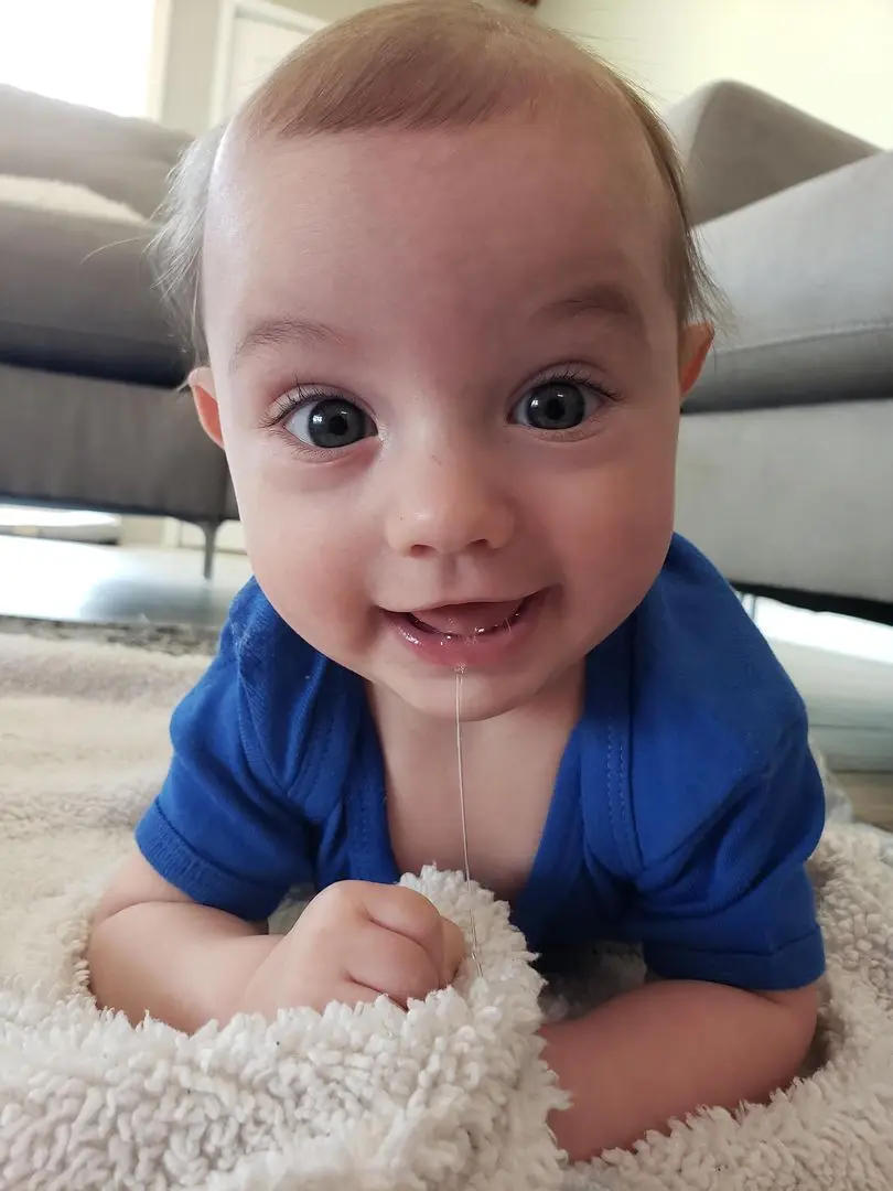 A smiling baby with light brown hair and a blue shirt lying on a white fluffy rug with a string of drool.
