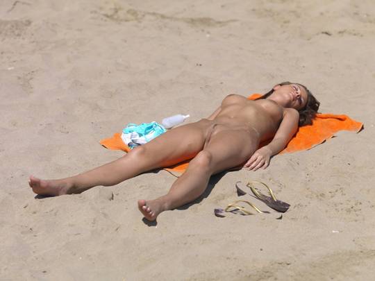 Slim brunette woman sunbathing nude on an orange towel at a naturist beach