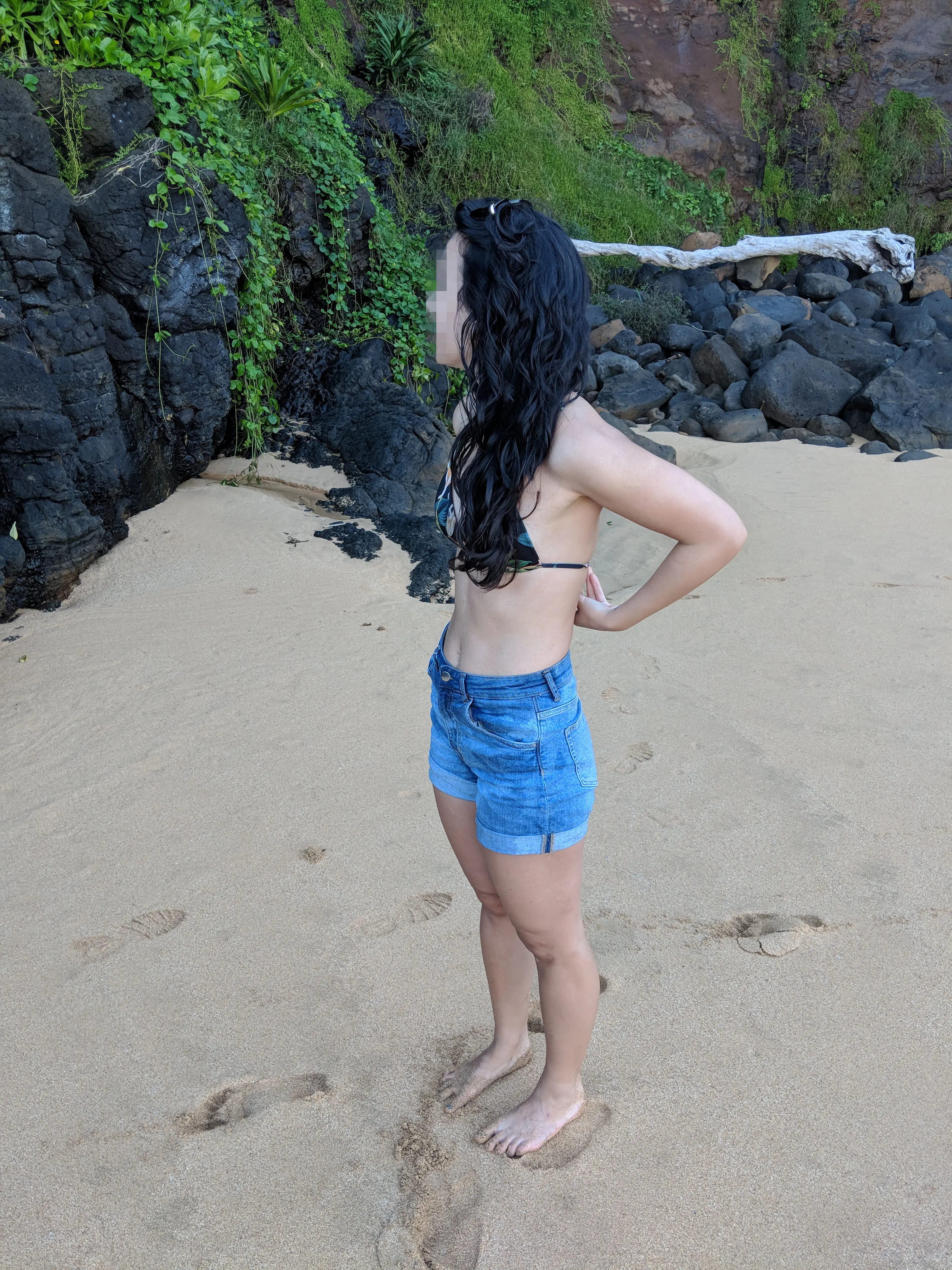 Slim Asian woman in denim shorts and a bikini top undressing to go topless on a Hawaii beach
