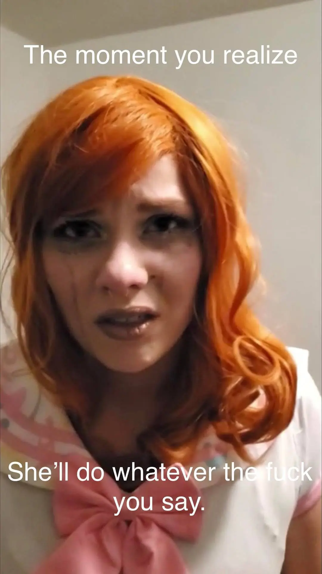A slim redhead woman in a white shirt and pink bow with a distressed expression, a dumbdoll.