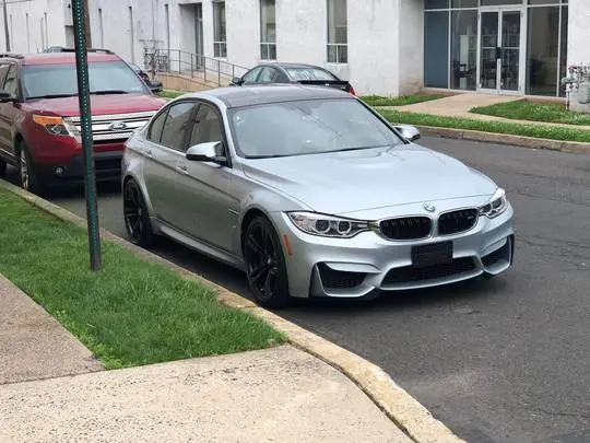 Aggressive silver BMW F80 sedan parked on an outdoor street next to a red Ford SUV and a white building.