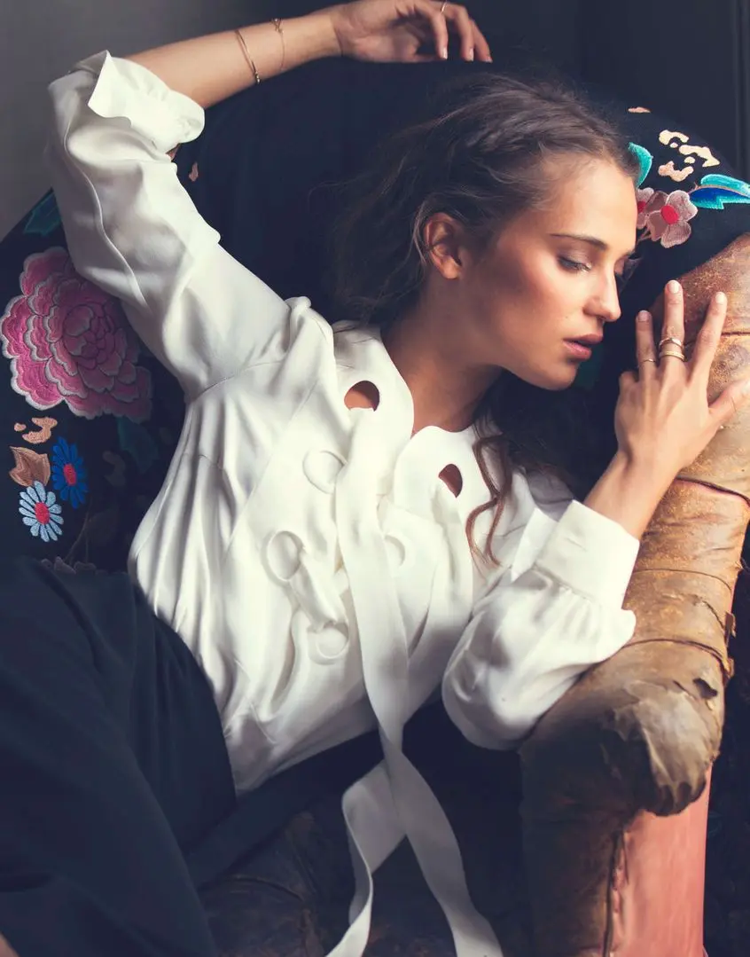 Alicia Vikander posing in a white suit, black dress, and floral gowns with a thigh tattoo