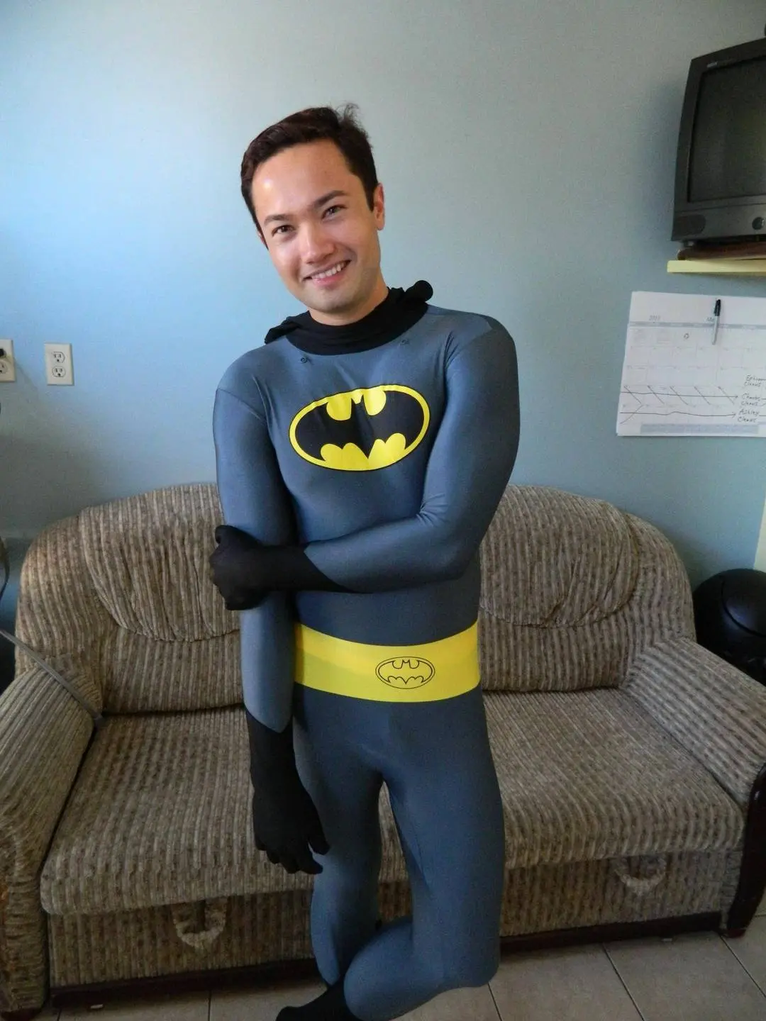 Slim Asian man in a grey and yellow Batman spandex bodysuit and black gloves smiling in a living room