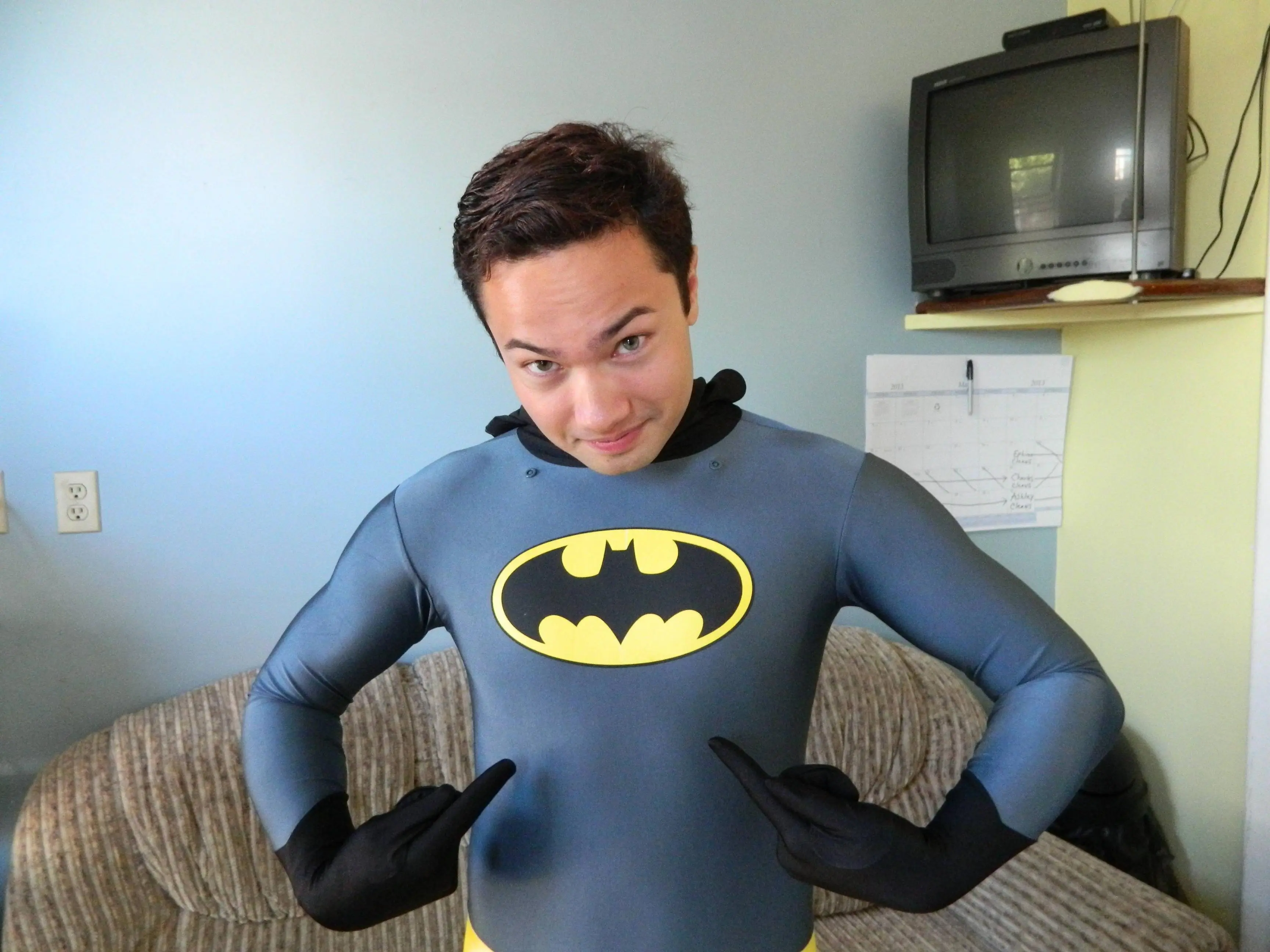 Slim Asian man in a grey and yellow Batman spandex bodysuit and black gloves smiling in a living room