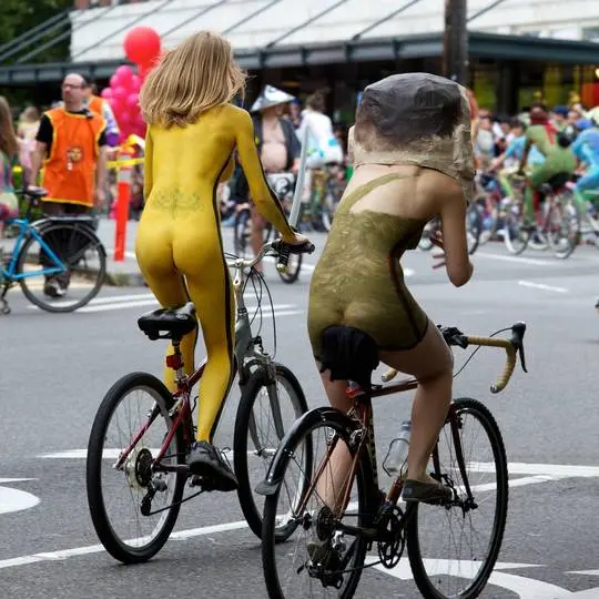 Slim brunette woman with yellow body paint riding a bicycle at a street fair, topless and nude