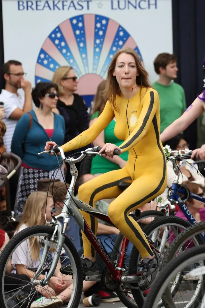 Slim brunette woman with yellow body paint riding a bicycle at a street fair, topless and nude