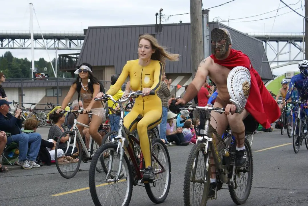 Slim brunette woman with yellow body paint riding a bicycle at a street fair, topless and nude