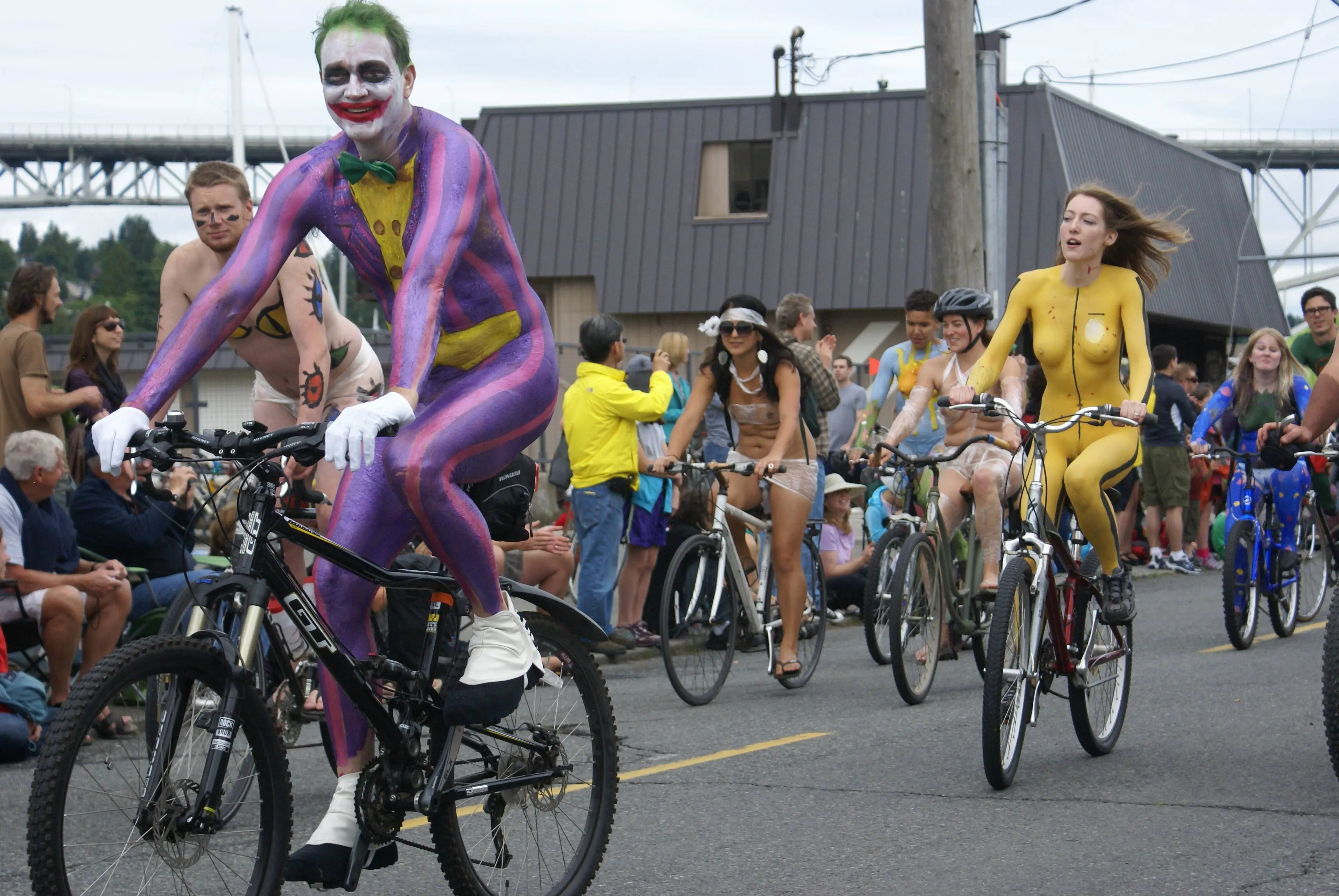 Slim brunette woman with yellow body paint riding a bicycle at a street fair, topless and nude
