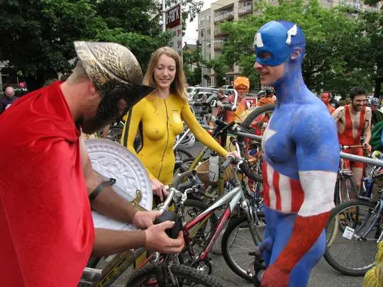 Slim brunette woman with yellow body paint riding a bicycle at a street fair, topless and nude