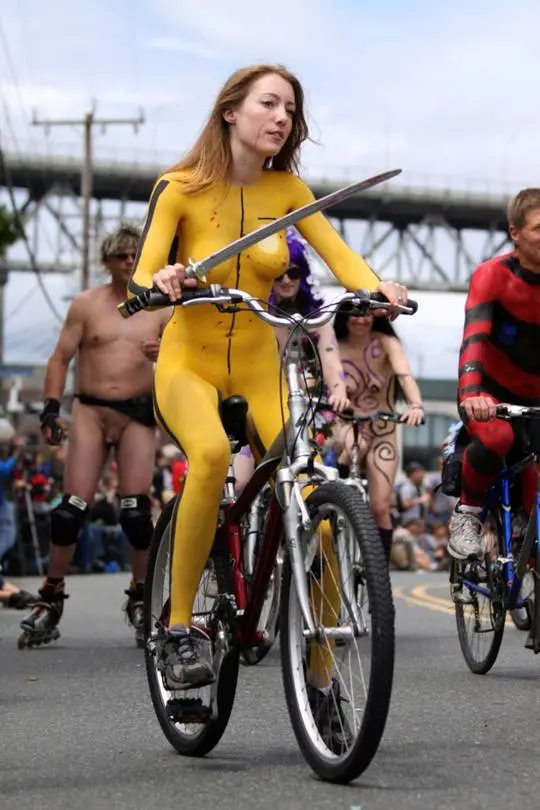 Slim brunette woman with yellow body paint riding a bicycle at a street fair, topless and nude