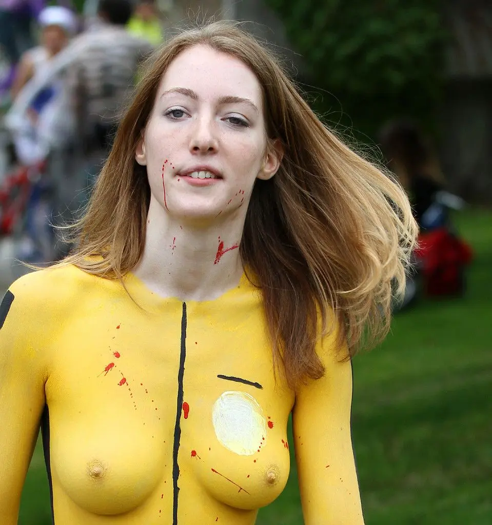 Slim brunette woman with yellow body paint riding a bicycle at a street fair, topless and nude