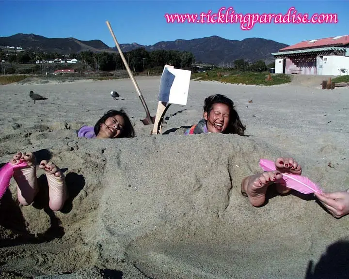 Asian women tickling feet and toes with a pink feather while buried in the sand at the beach