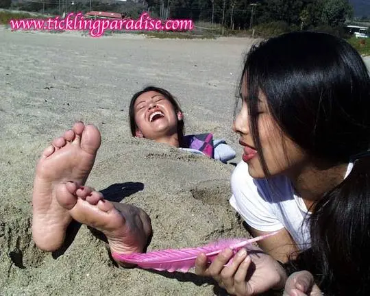 Asian women tickling feet and toes with a pink feather while buried in the sand at the beach