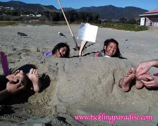 Asian women tickling feet and toes with a pink feather while buried in the sand at the beach