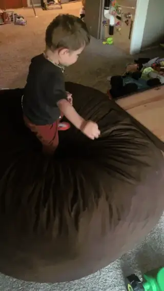 A light-skinned child in a grey t-shirt and reddish-brown pants falling off a brown beanbag chair in a living room.