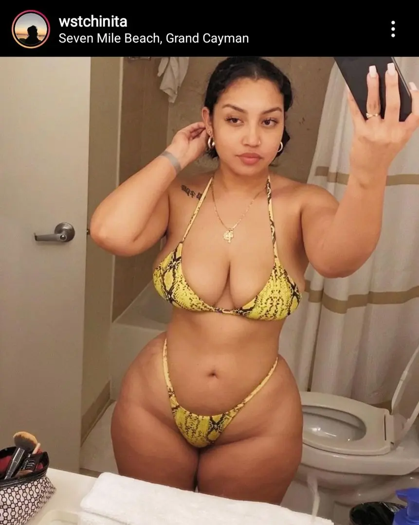 Curvy woman with tanned skin in a yellow snake-print bikini taking a mirror selfie in a bathroom