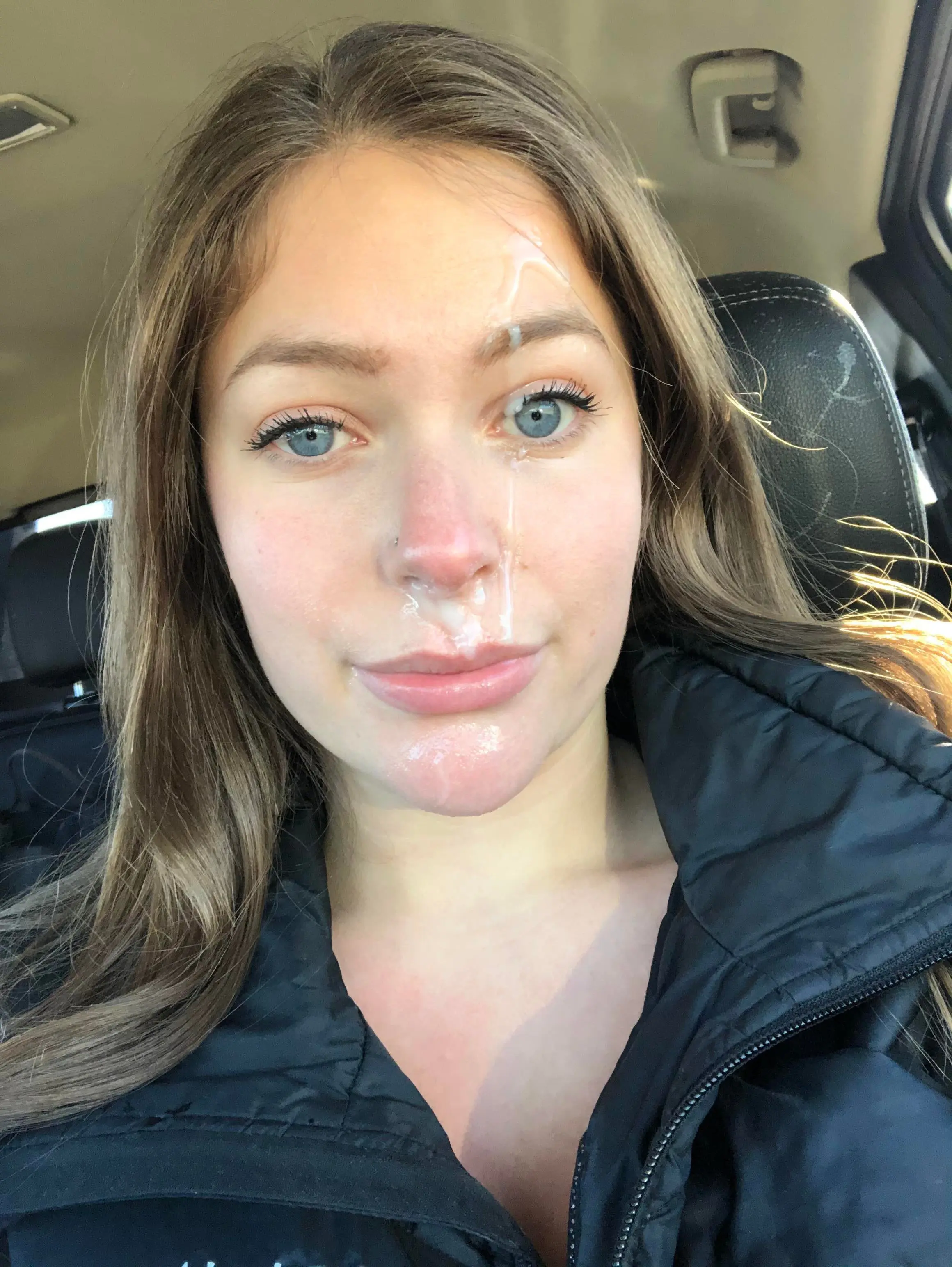 Slim white woman with long brown hair and a black puffer jacket in a car with a facial cumshot selfie