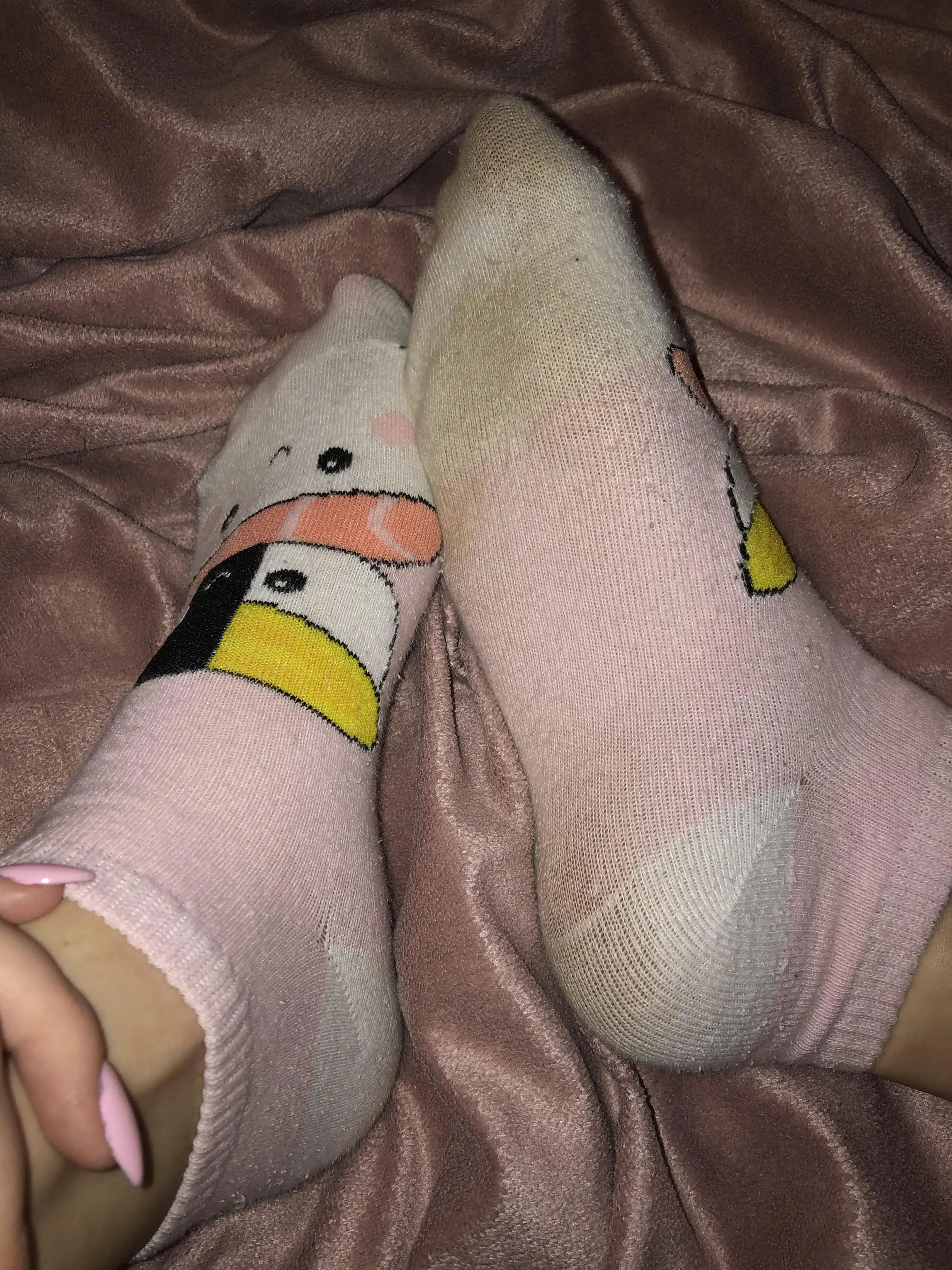 Close-up of a slim woman's feet in pink cartoon socks resting on pink satin sheets with pink nail polish.