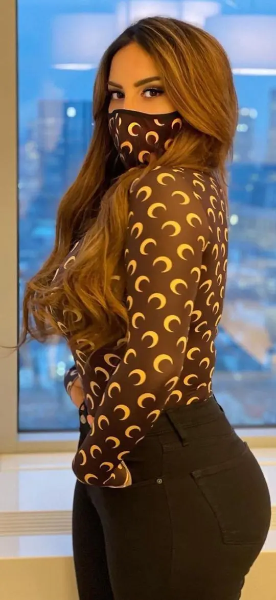 Enissa Amani wearing a brown crescent moon print top and face mask posing by a city skyline window
