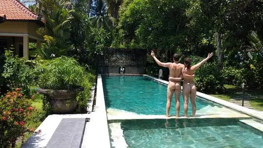 Nudist couple standing with arms raised by a swimming pool at a naturist resort.