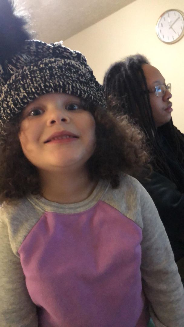 A child with curly brown hair in a black and white knit hat and purple shirt making a funny face