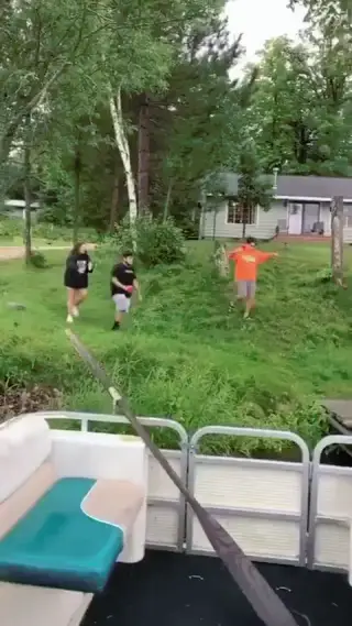 Three friends in hoodies and shorts running and tumbling on a grassy slope toward a wooden dock by a lake.