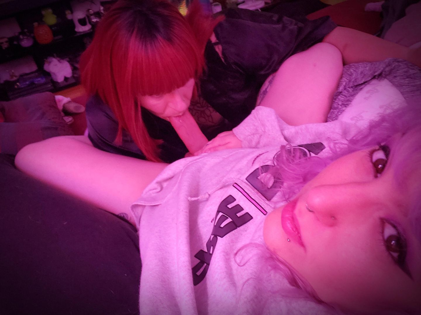 Trans woman with red hair and tattoos receiving oral sex while wearing a harness and vibrator, taking a selfie