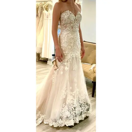 A person with fair skin wearing a strapless white lace wedding dress with a subtle blush underlay in a bridal boutique.