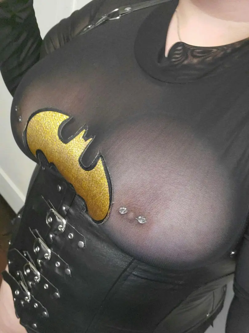 Busty white goth girl in a sheer black Batman top and black leather corset with nipple piercings