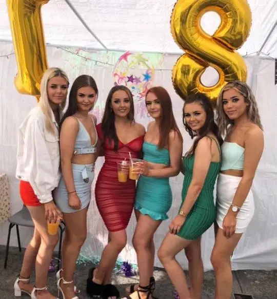Six ranked girls posing at a birthday party in colorful dresses and skirts under a white tent with gold balloons.