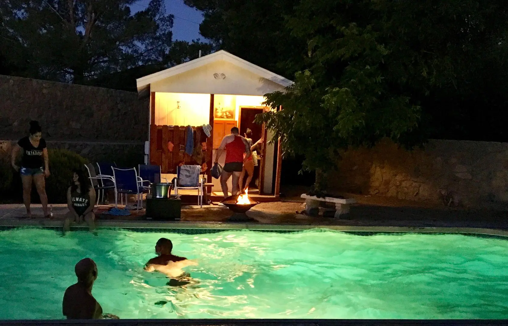A group of friends relaxing by a swimming pool and fire pit at a sauna party near a small white building.