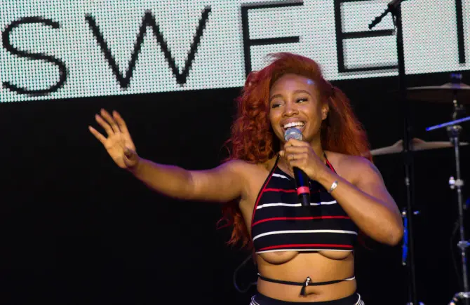 SZA performing on stage, a curvy black woman with long red curly hair in a striped crop top and green shorts