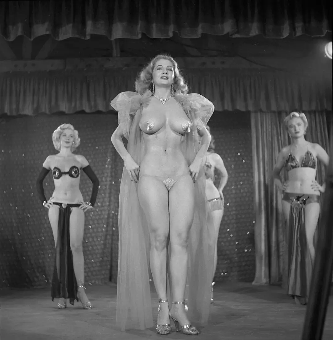 Tempest Storm and two other blonde women in revealing 1950s pinup costumes posing on a stage with a glittery backdrop