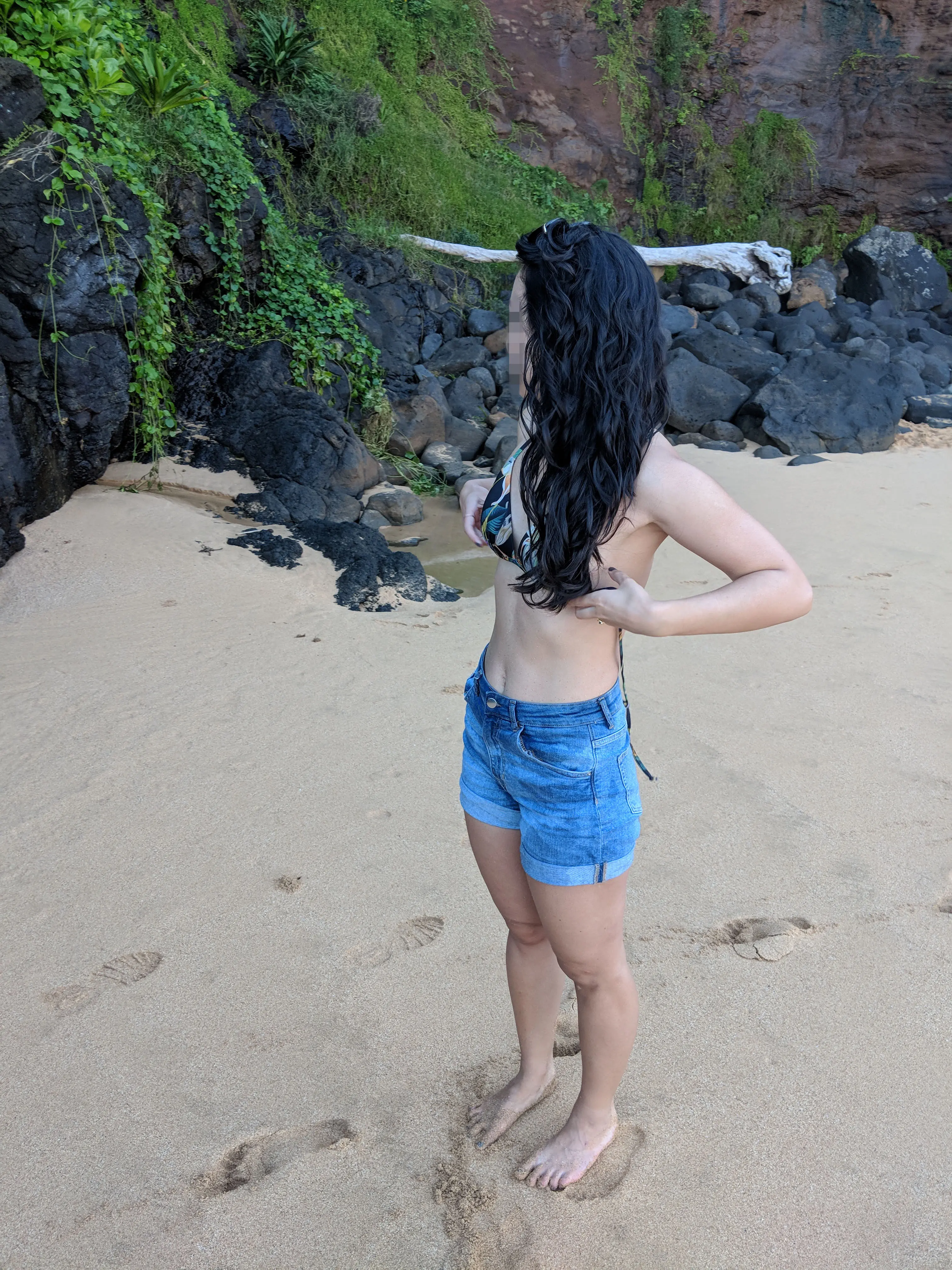 Slim Asian woman in denim shorts and a bikini top undressing to go topless on a Hawaii beach