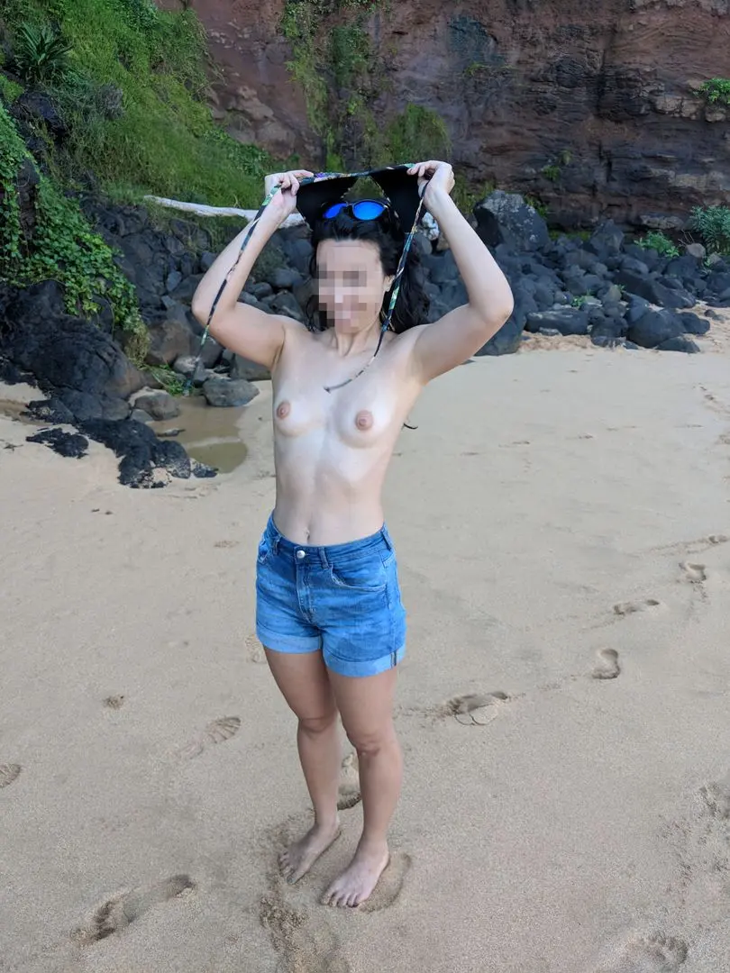 Slim Asian woman in denim shorts and a bikini top undressing to go topless on a Hawaii beach