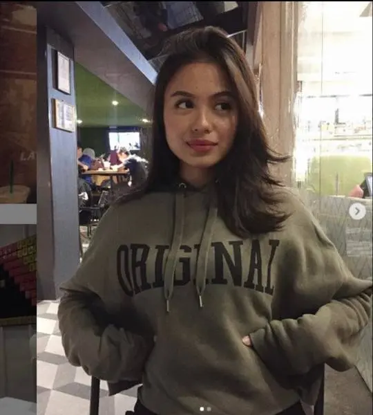 Slim tanned Asian woman in an olive green ORIGINAL hoodie posing in a cafe