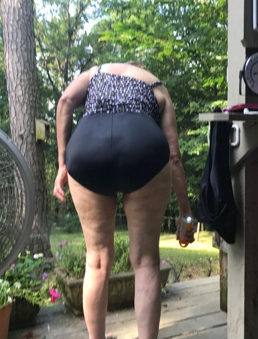 Curvy mature woman in a patterned top and black booty shorts, bent over from a back view on an outdoor deck.
