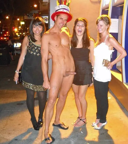 CFNM public nudity: a naked athletic white man with a party hat and erection posing with three slim women on a city sidewalk