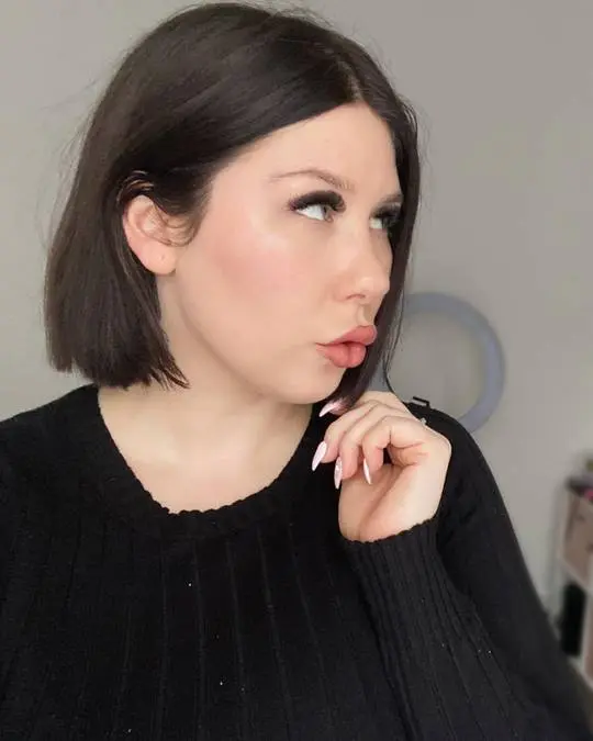 Findom woman with a short dark brown bob wearing a black ribbed sweater posing for a selfie.